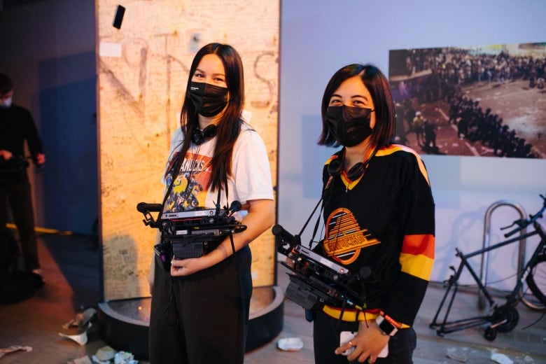 Filmmakers Kat Jayme and Asia Youngman stand together holding their cameras and wearing Vancouver Canucks shirts, masks, and headphones.